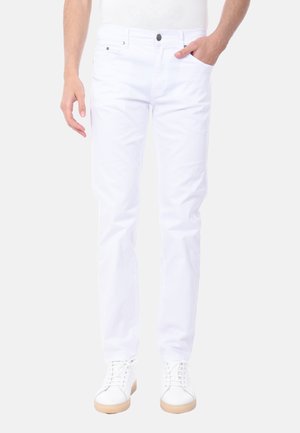 Uomo che indossa jeans bianchi slim-fit e sneakers bianche, con una mano in tasca, in piedi davanti a uno sfondo chiaro e uniforme.