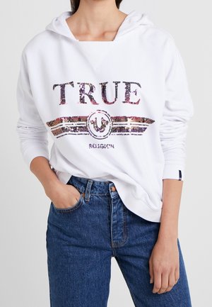 Biała bluza z kapturem z napisem "TRUE" i logo z cekinów, noszona wsunięta w niebieskie jeansy z wysokim stanem, lewa ręka modelki w kieszeni.