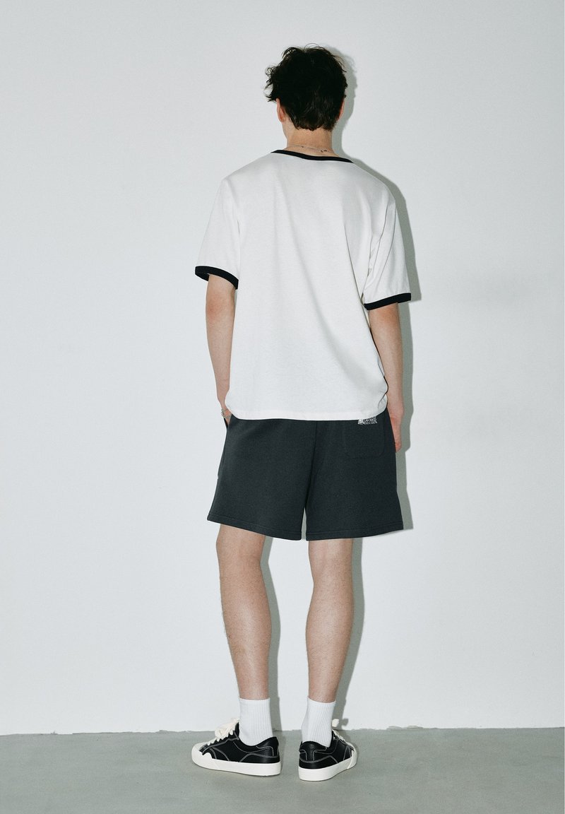 Jeune personne debout face à un mur blanc portant un t-shirt blanc avec un liseré noir, un short noir, des chaussettes blanches et des baskets noires.