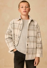 Chaqueta de franela a cuadros en crema y gris, con botones negros, llevando una camisa gris debajo y jeans negros. Textura suave y corte relajado.