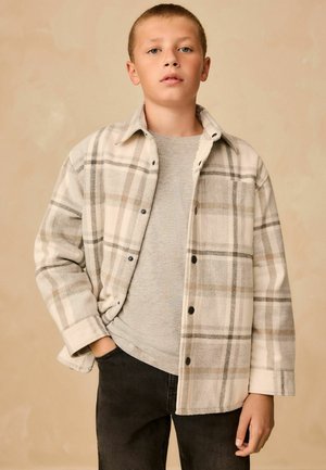 Rutet flanneljakke i creme og grå, med sorte knapper, en grå skjorte under og sorte jeans. Glat tekstur og afslappet pasform.