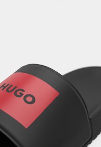 Melna slaidu sandale ar plašu siksnu, kurai ir sarkans kvadrāts un izteiksmīgi melns "HUGO" teksts.