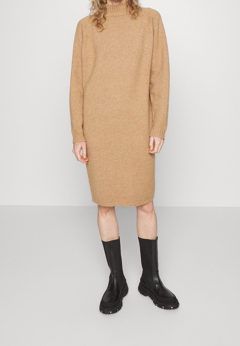 Personne portant une robe en tricot beige à manches longues et des bottes noires épaisses mi-mollet, debout sur un fond gris clair uni.
