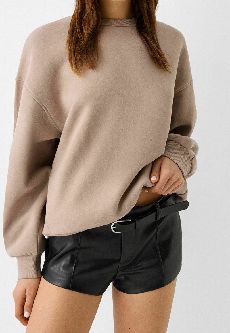 Personne portant un sweatshirt beige ample et un short en cuir noir avec une ceinture noire, debout devant un fond blanc uni.
