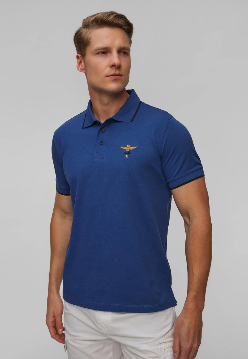 Polo blu con colletto, dotato di un emblema ricamato sulla parte sinistra del petto e accenti neri a contrasto lungo il colletto e le maniche.