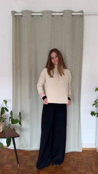 Jasnobeżowa bluza z zamkiem błyskawicznym i różowymi akcentami, w połączeniu z czarnymi szerokimi spodniami, na tle teksturowanej szarej zasłony.