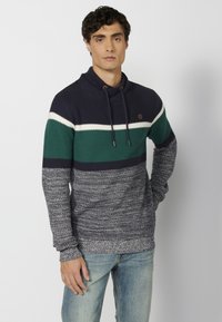 Sweter w paski w odcieniach granatu i zieleni z wysokim kołnierzem, sznurkiem i teksturowanym materiałem. Przerzedzone niebieskie dżinsy dopełniają całość.