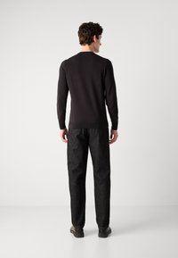 Pulover negru cu guler rotund, design ajustat, combinat cu pantaloni texturati de culoare gri închis. Bărbatul stă cu spatele, purtând pantofi negri.
