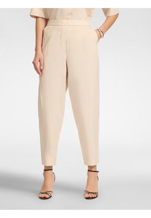 Donna che indossa pantaloni beige a vita alta e stretti con tasche e sandali neri con tacco e cinturini, in piedi con una mano in tasca.