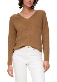 Pull en maille côtière marron avec un décolleté en V et des manches longues, associé à un jean blanc. Le matériau présente un motif texturé sur toute sa surface.
