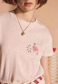 Biele bavlnené tričko s ružovým potlačeným obrázkom homára, hviezd a čiernych detailov. Nosí sa so zlatým retiazkou a perlovým príveskom.