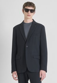 Crni strukturirani blejzer s dva prednja džepa i jednim dugmetom. Nosi se preko sive majice. Model nosi uglaste crne sunčane naočale.