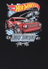 Černé tričko s grafikou červeného auta Hot Wheels s velkými pneumatikami, pozadí s mřížkovým vzorem a textem "NIGHT SHIFTER" v modré barvě.
