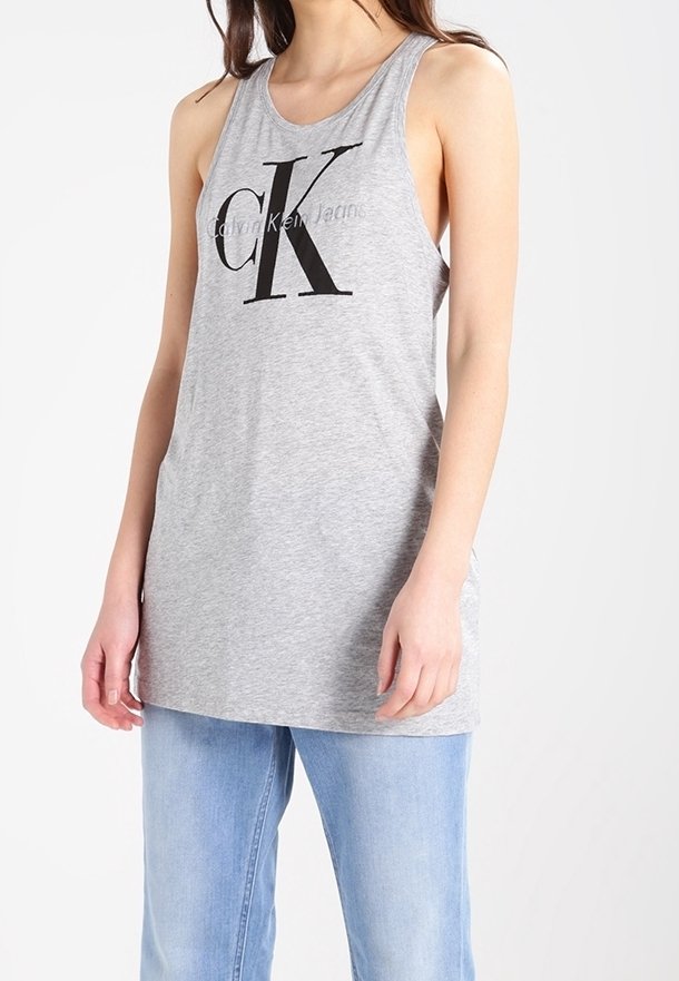 Femme portant un débardeur gris clair Calvin Klein sans manches avec un grand logo noir "CK" et un jean bleu clair, debout avec les bras détendus.