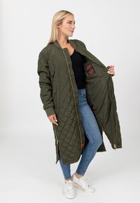 Zielono-oliwkowy, pikowany długi płaszcz z zamkiem błyskawicznym, prążkowanymi mankietami i bocznymi kieszeniami. Podszewka w kolorze pomarańczowym widoczna wewnątrz.