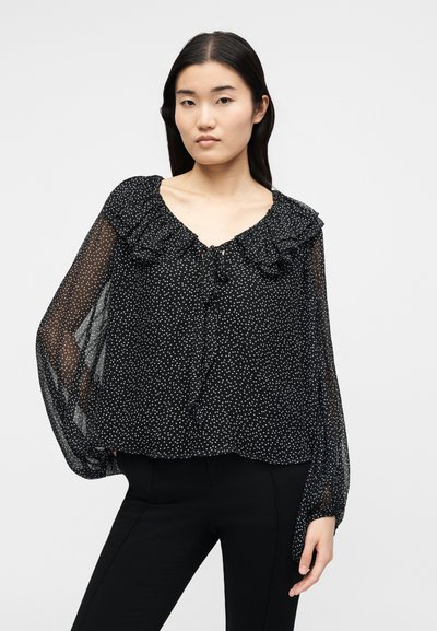 Νεαρή γυναίκα φοράει μαύρο διάφανο μπλουζάκι με λευκές πουά, βολάν στη λαιμόκοψη και μακριά φουσκωτά μανίκια, συνδυασμένο με μαύρο παντελόνι.