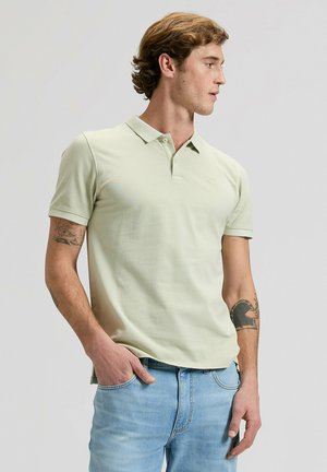 Giovane uomo con tatuaggi indossa una polo verde chiaro e jeans azzurri, guarda verso destra con una mano in tasca su sfondo neutro.