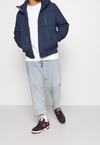 Chaqueta acolchada navy con capucha, bolsillos con cremallera, camiseta blanca, jeans de denim claros y zapatillas negras con acentos rojos.