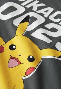 Geltona Pikachu grafika ant pilkos medžiagos su balta išklotine, apimančia juodus akcentus, blizgiomis akimis ir besišypsančia išraiška.