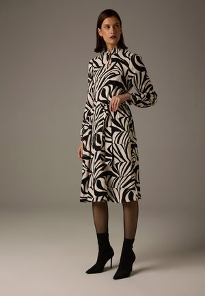 Schwarz-weiß gemustertes Kleid mit hohem Kragen, langen Ärmeln, gebundener Taille und einem fließenden Rock. Kombiniert mit schwarzen spitzen Stiefeletten und Netzstrumpfhosen.