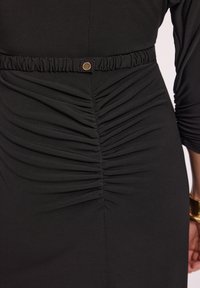 Czarna dopasowana sukienka z marszczeniami wzdłuż talii i elastycznym pasem. Złoty guzik ozdobny na zgromadzonym materiale.