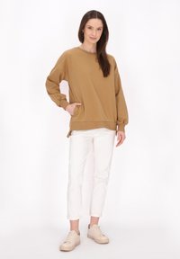 Felpa marrone oversize con scollatura a girocollo e spacchi laterali, abbinata a pantaloni bianchi arrotolati e sneakers beige. Tessuto morbido.