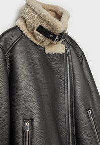 Schwarze Lederjacke mit cremefarbenem Schaffellkragen und schwarzem Schnallenriemen, mit vorderen und seitlichen silbernen Reißverschlüssen vor weißem Hintergrund.