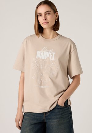 Jeune femme aux cheveux courts et bruns portant un t-shirt beige avec un motif de fleur blanche et le texte « flower MARKET », associé à un jean bleu, main dans la poche.