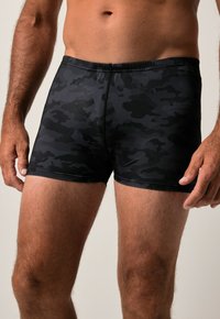 Torse masculin et cuisses supérieures portant un maillot de bain ajusté à motifs camouflage noir et gris foncé, mains reposant sur les côtés.