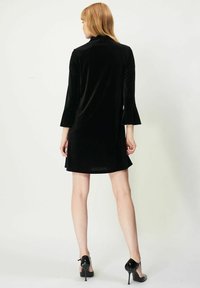 Svart sammetsklänning med utsvängda, trekvartslånga ärmar; knälång längd med en slät yta; bärs med svarta högklackade skor.