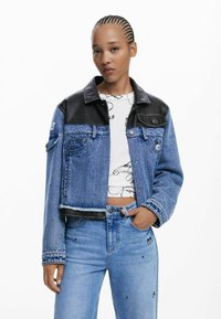 Giacca di jeans con yoke in pelle nera, design cropped, colore azzurro chiaro, due tasche frontali e orlo sfilacciato. Indossata sopra una maglietta bianca con grafica.
