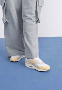 Sapatilhas Nike brancas e bege com bico arredondado, parte superior em malha e logótipo na lateral, combinadas com calças de fato cinzentas e folgadas.