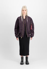 Sieviete ar garām blondām matēm, ģērbusies tumši purpura bomber jakā, melnā midi kleitā un melnās potīšu zābakos, stāv uz vienkārša balta fona.