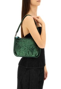 Borsa a tracolla verde con paillettes, dal texture liscia e forma strutturata. Presenta un manico corto e una chiusura con zip, indossata da un modello.