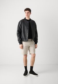 Black faux leather bomber jacket with ribbed cuffs, paired with beige shorts, black sneakers, and black socks. Model stands against a white background.