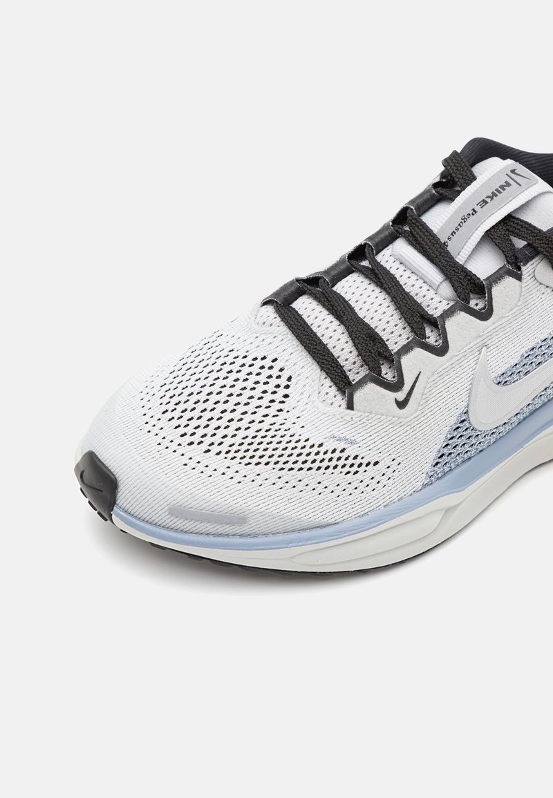 Scarpa sportiva bianca con tomaia in rete, lacci neri e dettagli azzurri chiari sulla suola. Presenta una punta arrotondata e una superficie testurizzata.