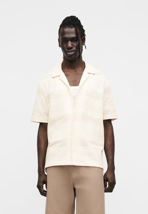 Kortærmet, cremefarvet strikket skjorte med vandrette striber og hulmønsterdetaljer, kombineret med lysebrune bukser.