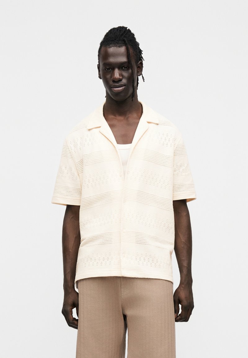 Κοντομάνικο πλεκτό πουκάμισο σε εκρού χρώμα με οριζόντιες ρίγες και διακοσμητικά ανοίγματα, συνδυασμένο με ανοιχτό καφέ παντελόνι.