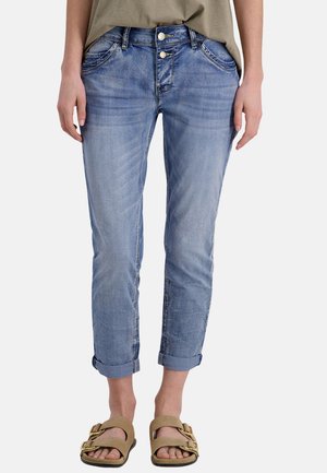 Hellblaue aufgerollte Jeans, getragen von einer stehenden Person, kombiniert mit beigefarbenen Sandalen mit großen Schnallen.
