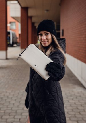 Cream leather wallet with a black zipper and logo, held in a gloved hand. Background features a brick building and cobblestone ground.