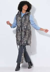Leopardenmuster Pufferweste mit Kapuze, Vorderreißverschluss und zwei Seitentaschen; getragen über einem gestreiften Hemd und kombiniert mit ausgestellten Jeans.