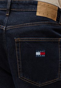 Σκούρα μπλε τζιν από denim με πορτοκαλί ραφές, με λογότυπο Tommy Jeans στην πίσω τσέπη και δερμάτινη ετικέτα της μάρκας στη μέση.