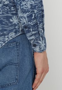 Blau gemustertes Hemd mit Palmenmotiv, mit Knopfmanschetten und lockerem Schnitt, kombiniert mit hellblauen Jeans.