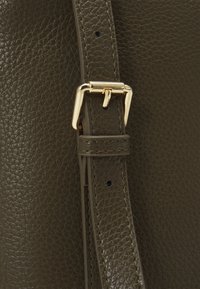 Olive green leather bag detail featuring textured surface, gold metal buckle, and adjustable strap with stitch accents.