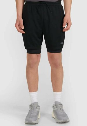 Mężczyzna w czarnych warstwowych sportowych szortach, białych skarpetkach typu crew oraz szarych butach do biegania, stojący na jednolitym tle.