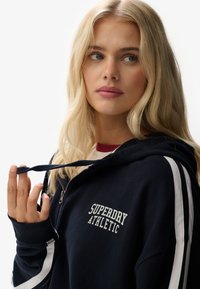 Sudadera con cremallera en color navy, con rayas blancas en los laterales, que presenta "Superdry Athletic" bordado en el lado izquierdo del pecho. Tela suave con un corte relajado.