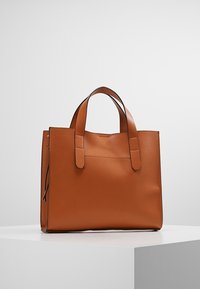 Sac fourre-tout en cuir beige avec double poignées, forme rectangulaire et design minimaliste, posé sur un socle blanc contre un fond uni.