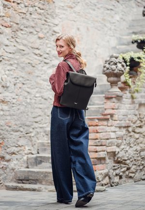 Černý kožený batoh s minimalistickým designem, vybavený překlopným víkem a nastavitelnými popruhy. Model má na sobě široké džíny.