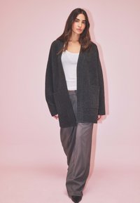 Žena s dlhými hnedými vlasmi, oblečená v tmavošedej kardigane, bielej blúzke a voľných šedých nohaviciach na svetloružovom pozadí.