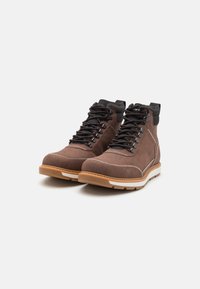 Botas de tobillo de ante marrón con acentos negros, que cuentan con un diseño de cordones, costuras blancas y una suela de goma color beige para mayor tracción.